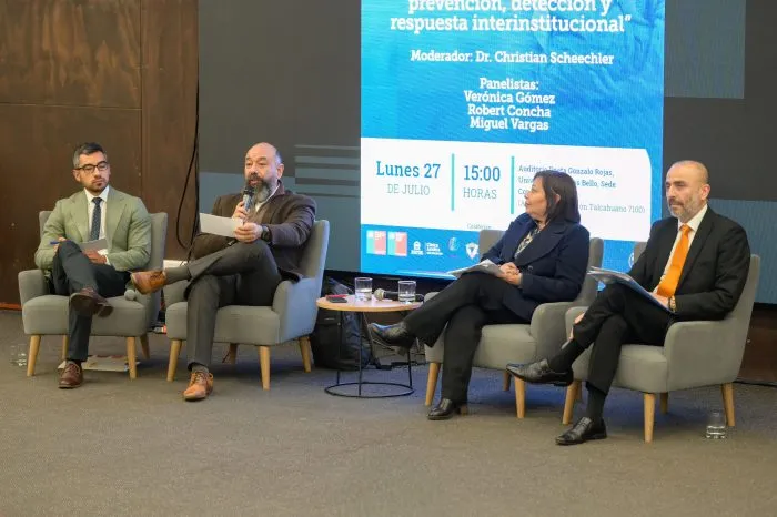 Four panelists seated on stage in gray chairs, one speaking into a microphone as the others listen; a large blue event banner is projected behind them.