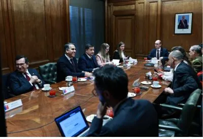 Group of professionals in a formal meeting around a wooden conference table with laptops and coffee cups.