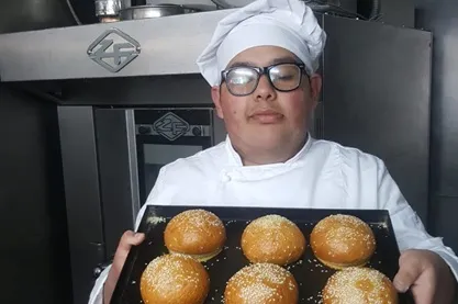 Chef in white uniform and glasses holds a baking tray with six sesame buns in a commercial kitchen, with an oven in the background.