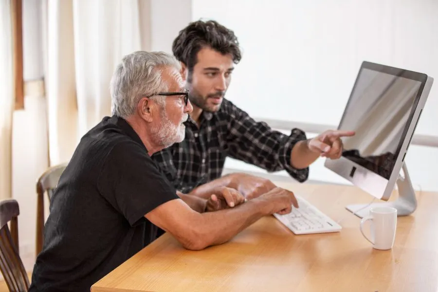 Two men collaborate at a desk, with the younger man guiding the older man as he points at a computer screen, keyboard in front of them.