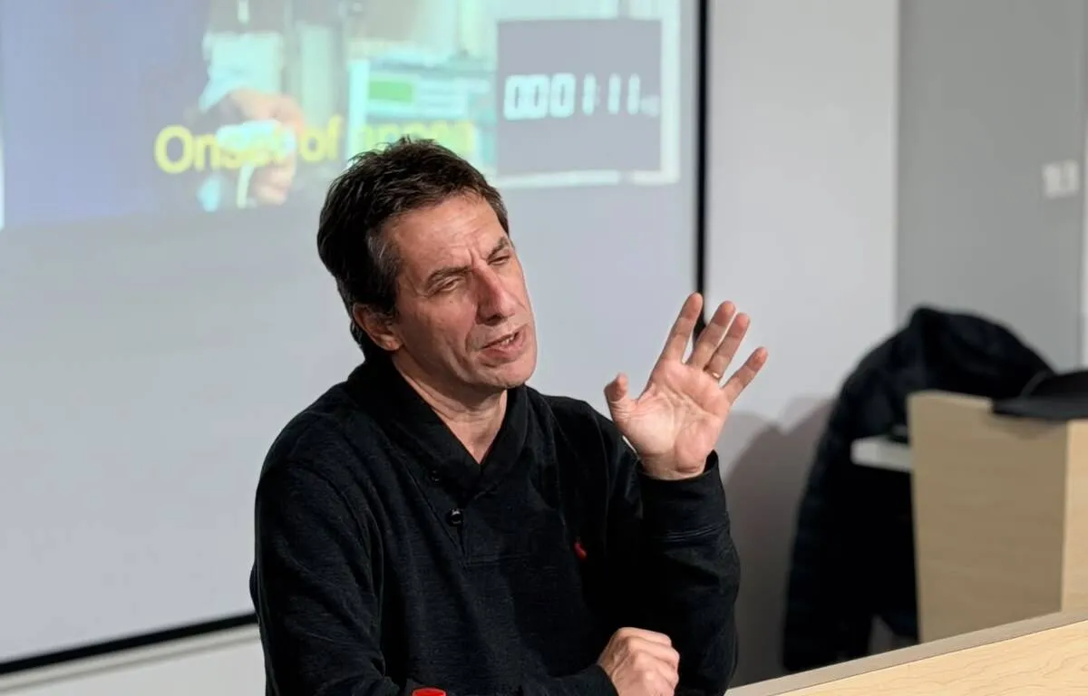 Man in a dark sweater speaks and gestures with his right hand in a lecture room, projection screen visible in the background.