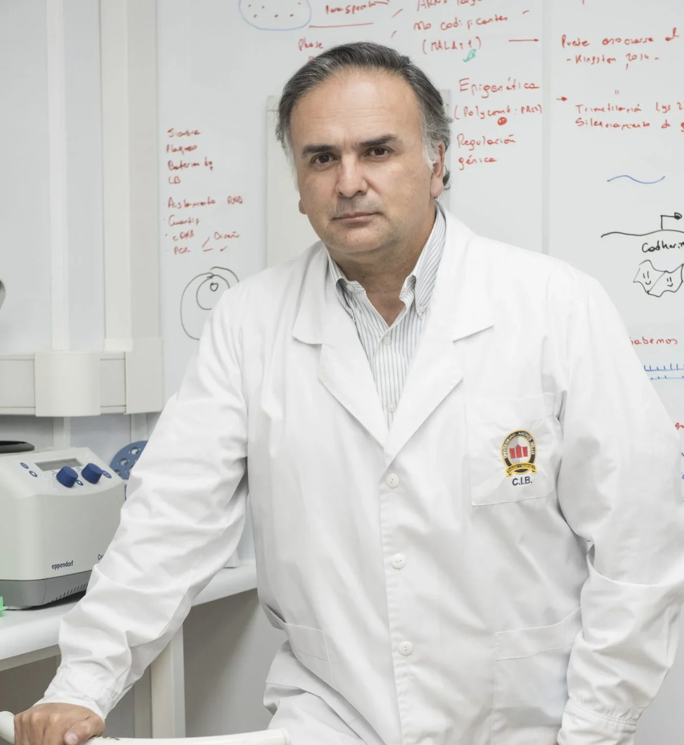 Male scientist in a white lab coat standing at a lab bench, with a whiteboard full of notes in the background.