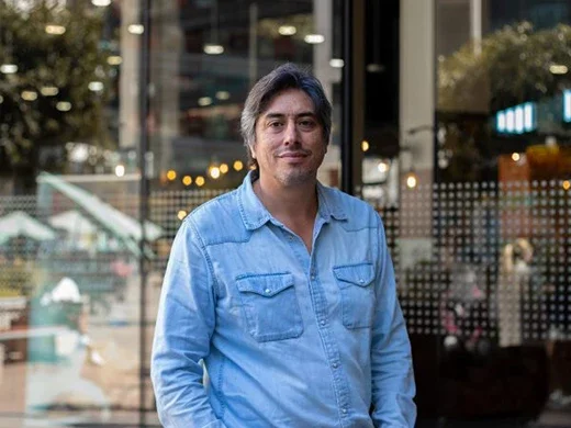 Man in a light blue denim shirt standing outside a storefront, facing the camera.