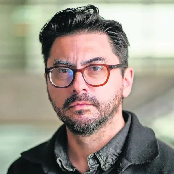 Portrait of a man with dark hair and beard wearing brown glasses, looking at the camera with a neutral expression in a blurred background.