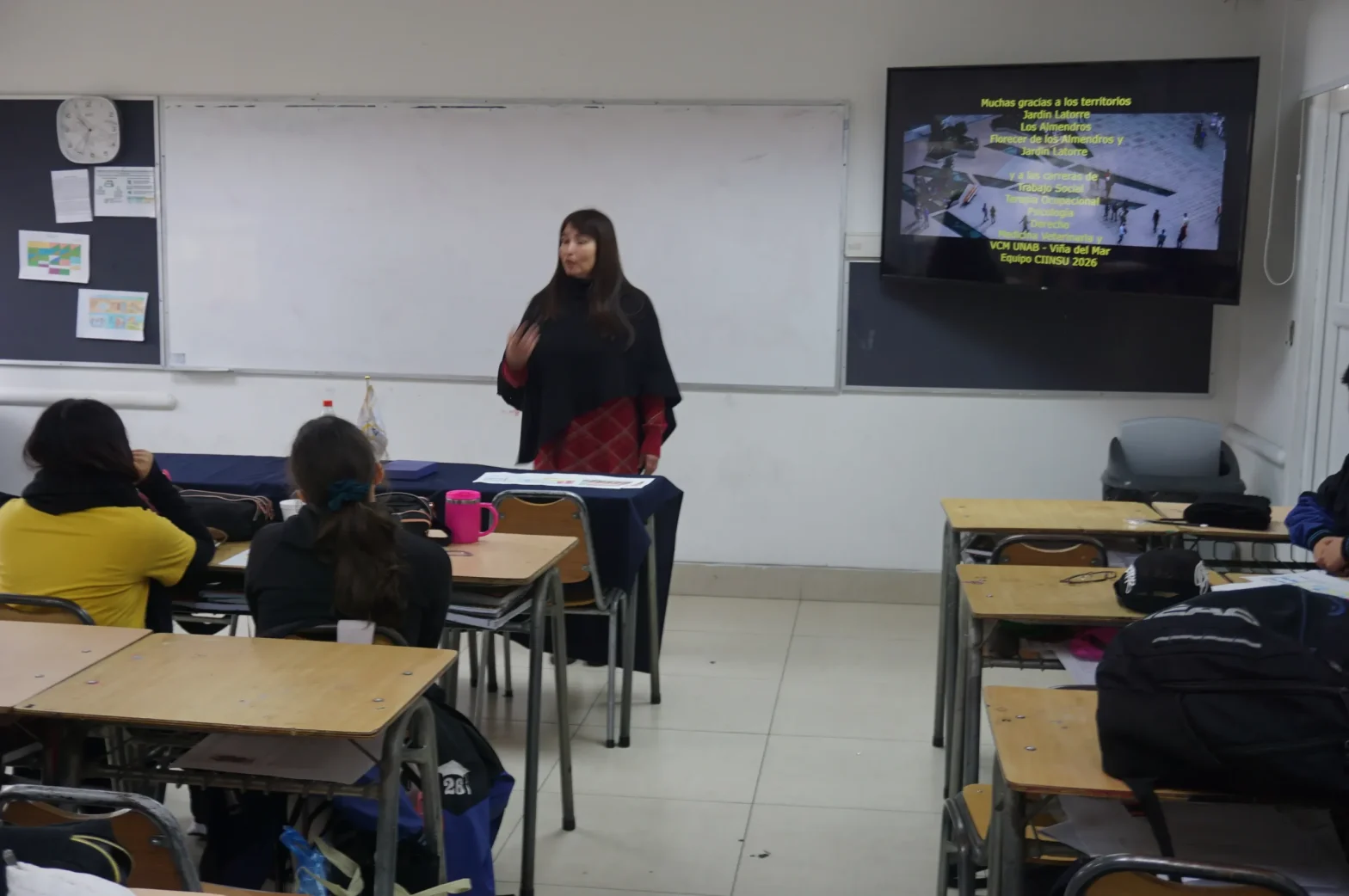 Teacher standing at the front of a classroom delivering a lecture; students sit at desks facing a whiteboard and a projector screen showing slides in Spanish.