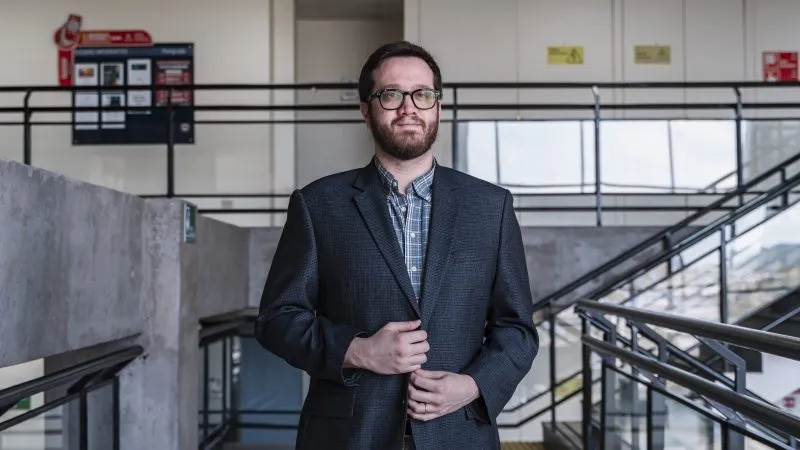 Man in a dark blazer and checkered shirt stands on a modern indoor stairwell, adjusting his jacket and looking at the camera.