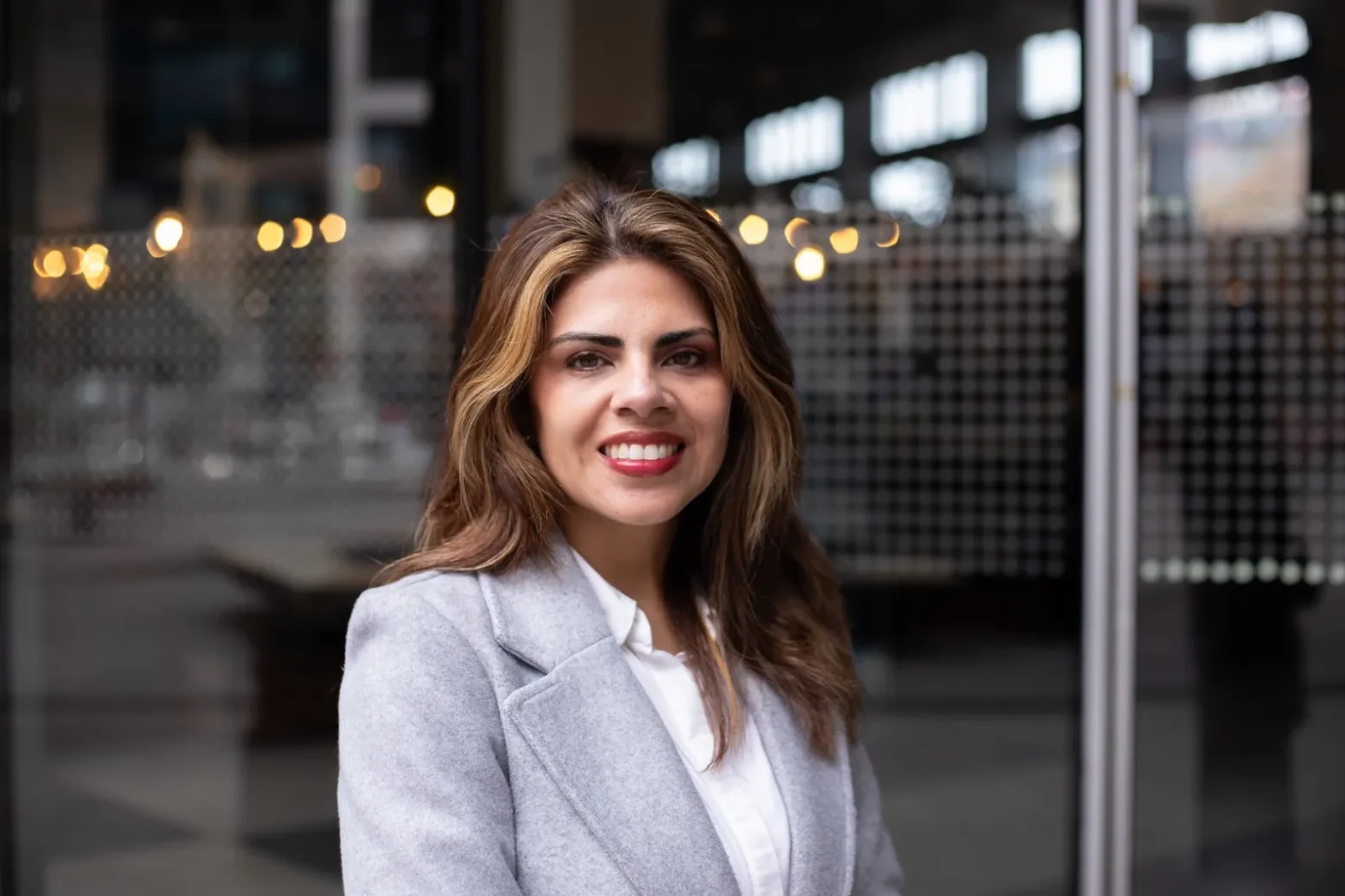 Smiling woman with light brown hair in a gray coat standing outside a glass building with blurred lights in the background.