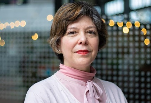 Portrait of a middle-aged woman with short brown hair wearing a light pink blouse, looking at the camera with a neutral expression.