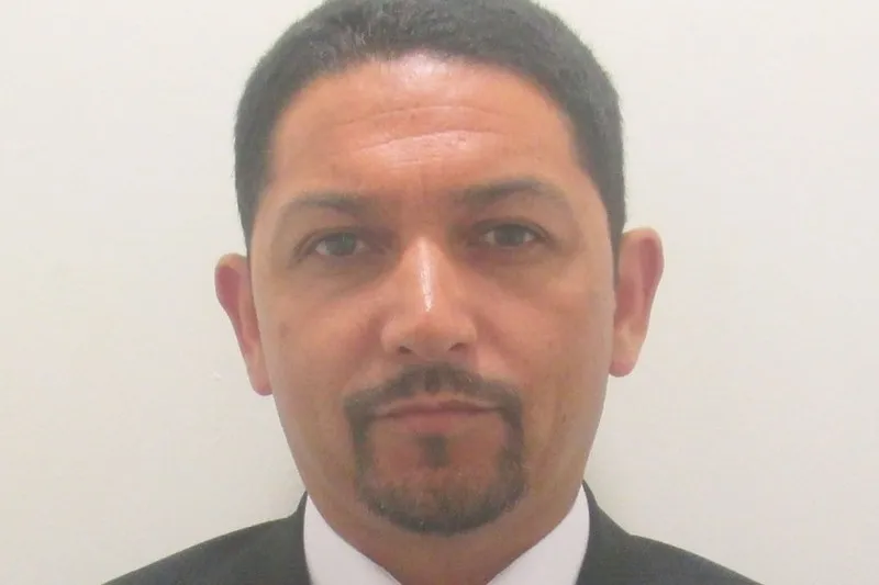 Man with short dark hair and goatee wearing a suit and white shirt, facing the camera with a neutral expression against a plain background.