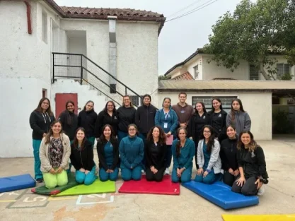 Group of about 20 people posing for a photo outdoors in front of a white building with stairs; several are seated on bright exercise mats.