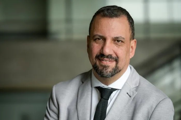 Professional headshot of a smiling man with short dark hair and a beard, wearing a light gray suit and black tie in a modern office setting.