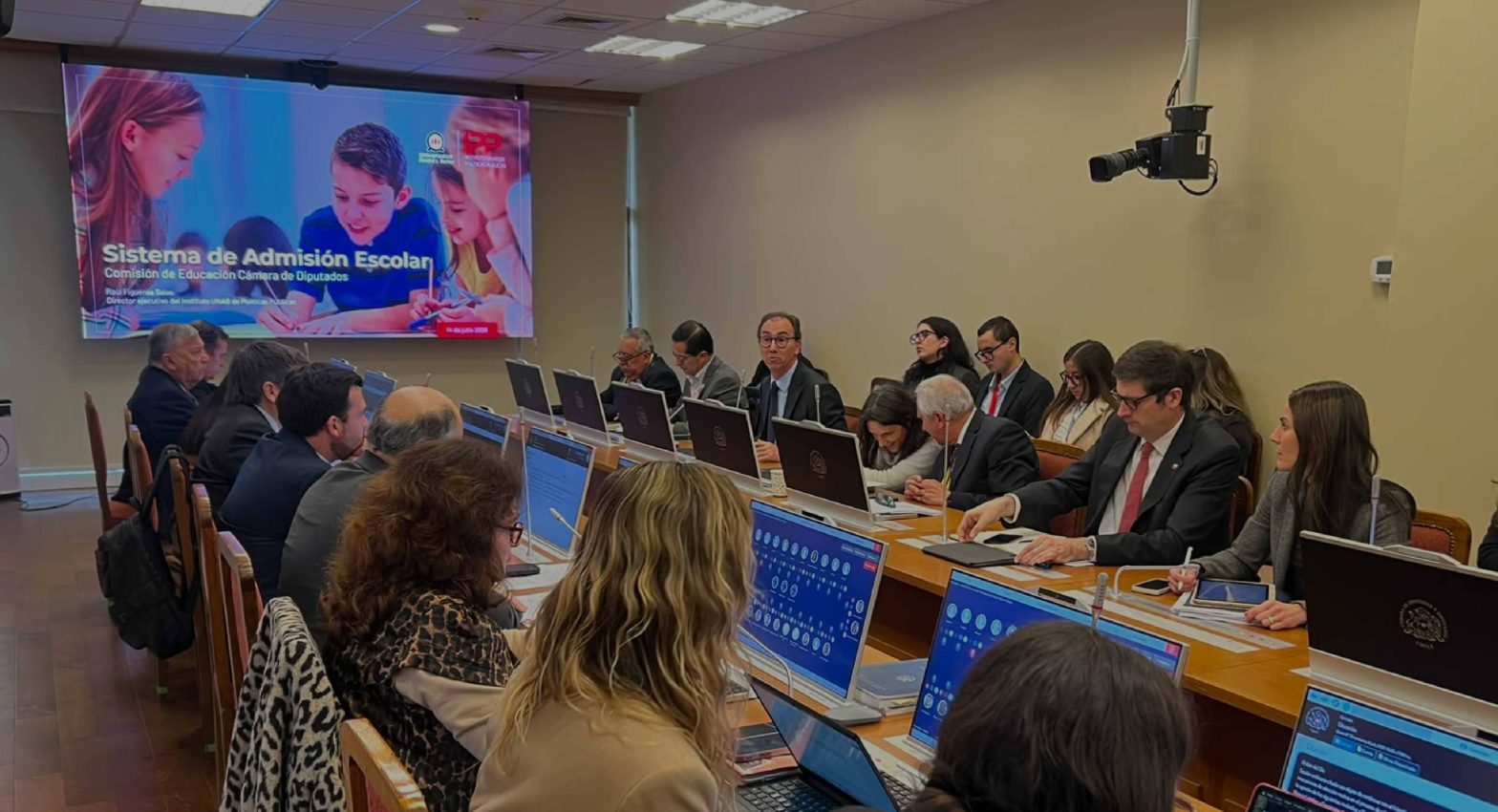 Group of professionals in a conference room seated along a long table with laptops and documents; a large banner reads 'Sistema de Admisión Escolar'.