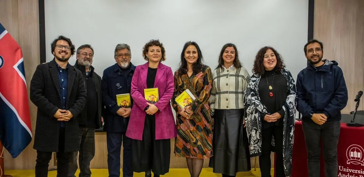 Group of eight diverse adults standing in a line on a stage, each holding a yellow booklet. The setting suggests a university event with a flag on the left and a table with a banner on the right.