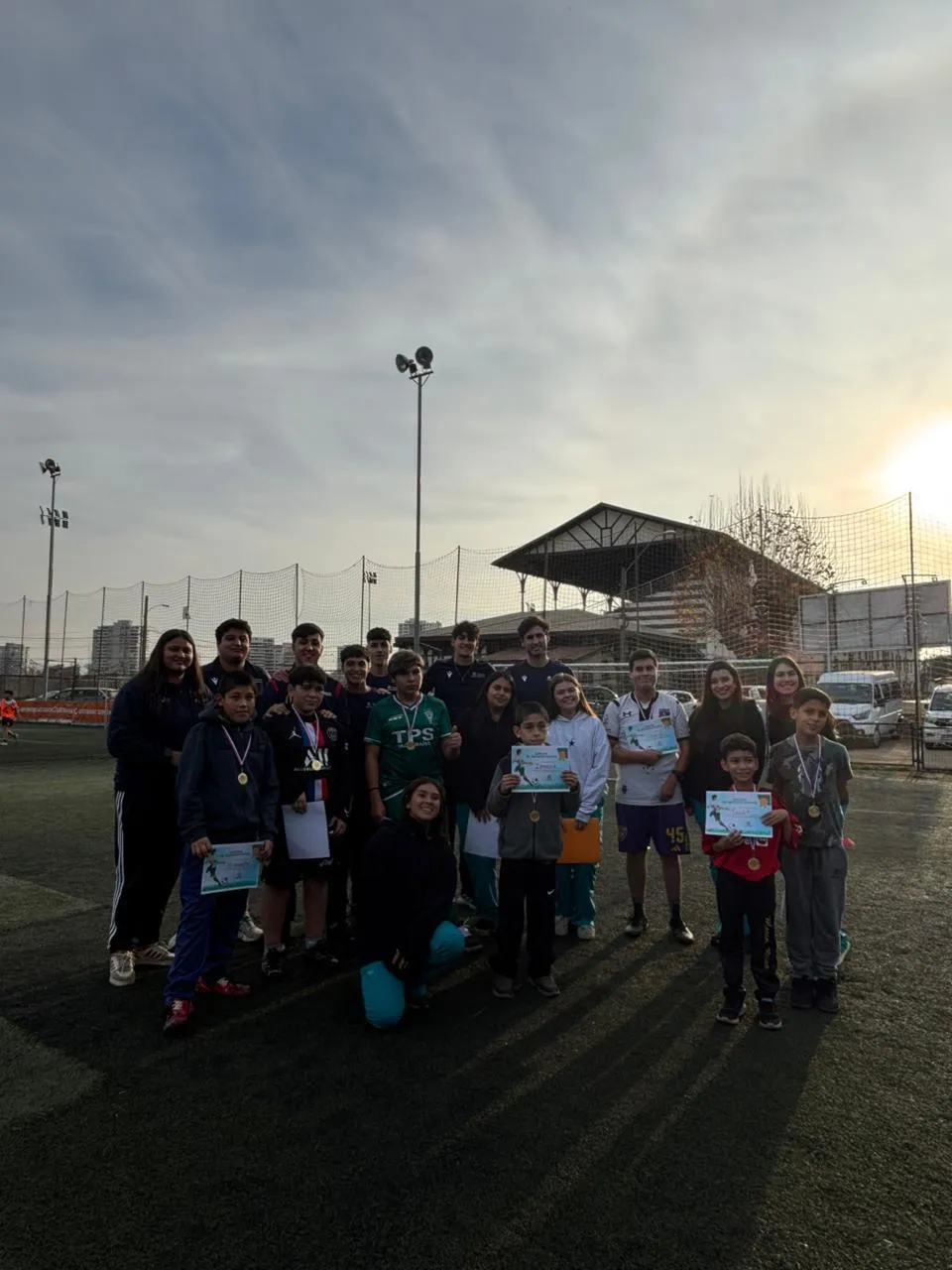 Group of kids and adults on a soccer field posing with certificates and medals at sunset.