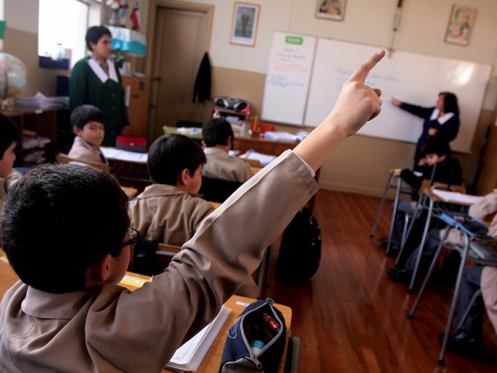 Student in a beige uniform raises a hand to answer during a classroom lesson with a teacher at the whiteboard in the background.