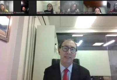 Man in a suit and red tie presenting in a video conference, with multiple participants shown in small thumbnail videos along the top.