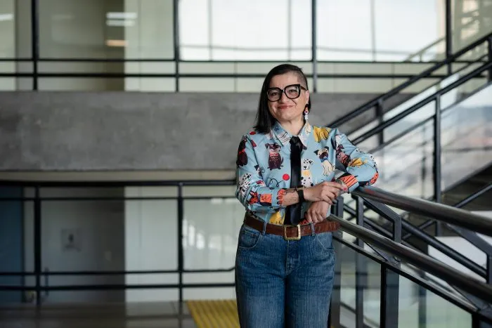 Woman with short dark hair and glasses standing in a modern atrium, leaning on a railing, wearing a colorful patterned shirt, blue jeans, and a brown belt.