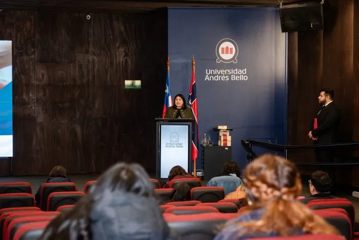 Female speaker at a podium in a lecture hall with Universidad Andrés Bello banner and flags nearby.