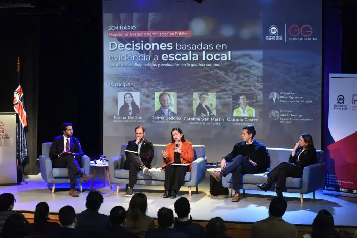 Panel of five speakers seated on stage during a seminar; a large screen behind shows the title 'Decisiones basadas en evidencia a escala local' and speaker portraits, with an audience in the foreground.