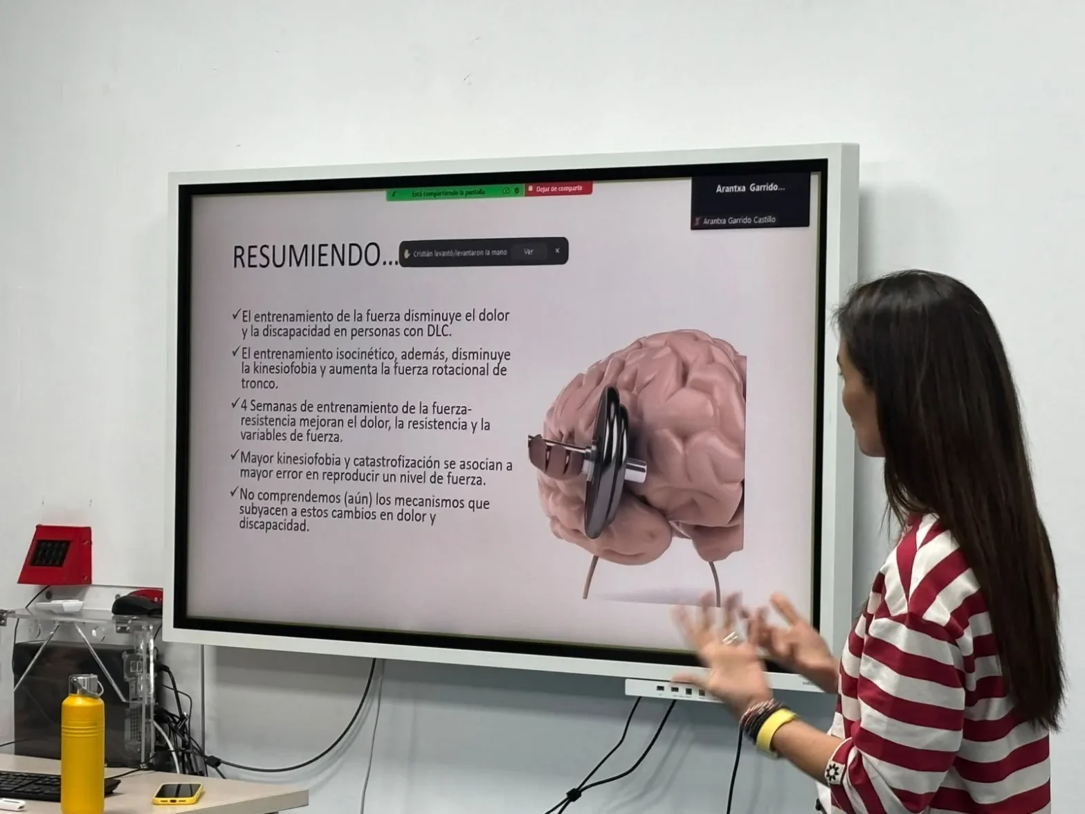 Presenter standing beside a large screen displaying a slide that starts with 'RESUMIENDO...' and shows a pink brain with a dumbbell, plus bullet points in Spanish.