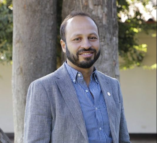 Man with a beard in a gray blazer and blue checkered shirt, smiling outdoors with trees behind him.