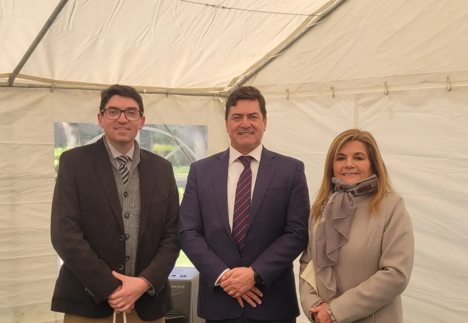 Three adults in business attire posing for a photo inside a white tent; two men in suits and a woman in a scarf smile at the camera.