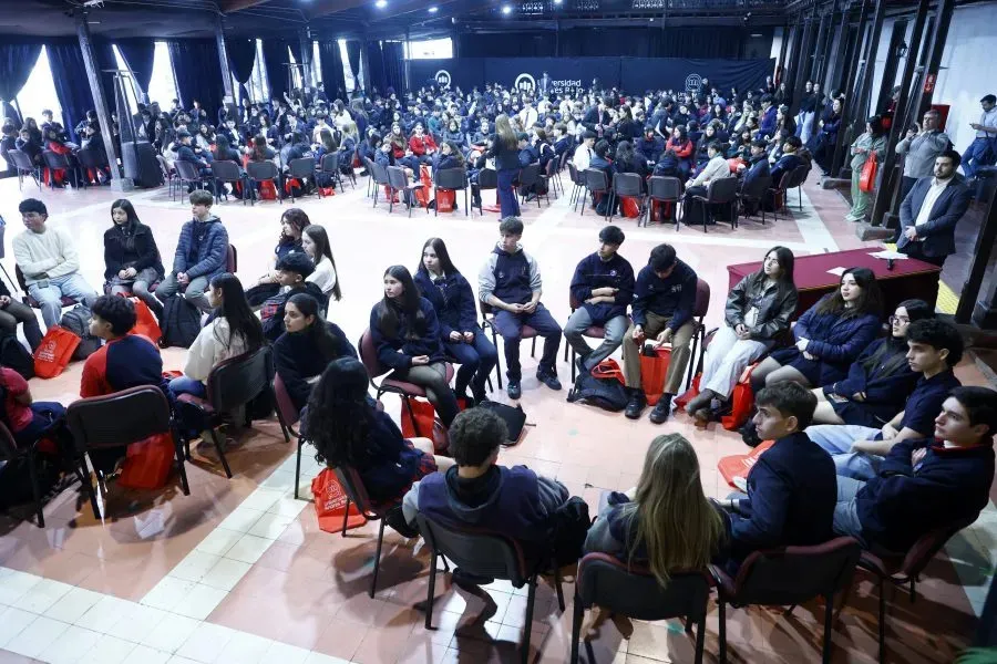 Large conference room filled with people seated in circular arrangements, several red bags on chairs, stage and backdrop visible in the background.