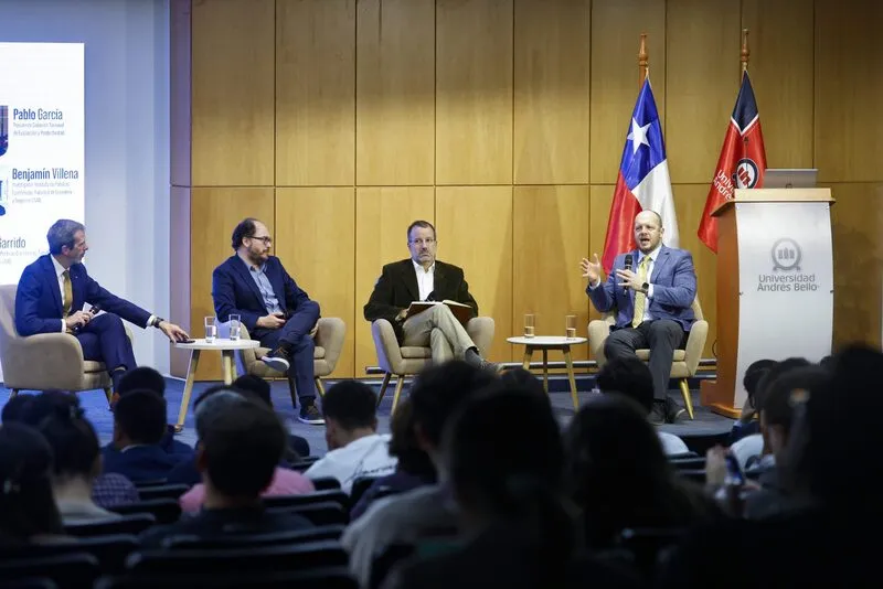 Panel discussion on stage with four men seated in armchairs; flags and podium to the right in a university venue.