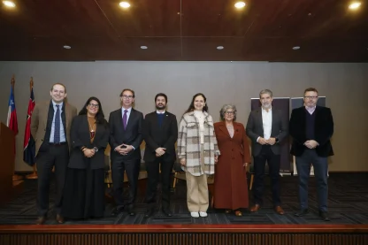 Eight adults in business attire pose on a stage with flag banners to the left, for a formal group portrait.