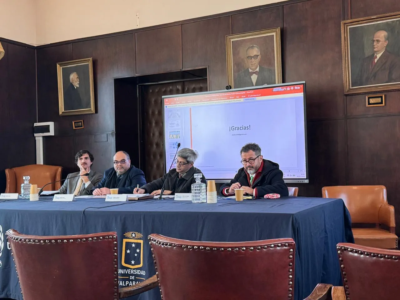 Panel discussion at a university with four men seated at a long table; a screen behind them displays '¡Gracias!'