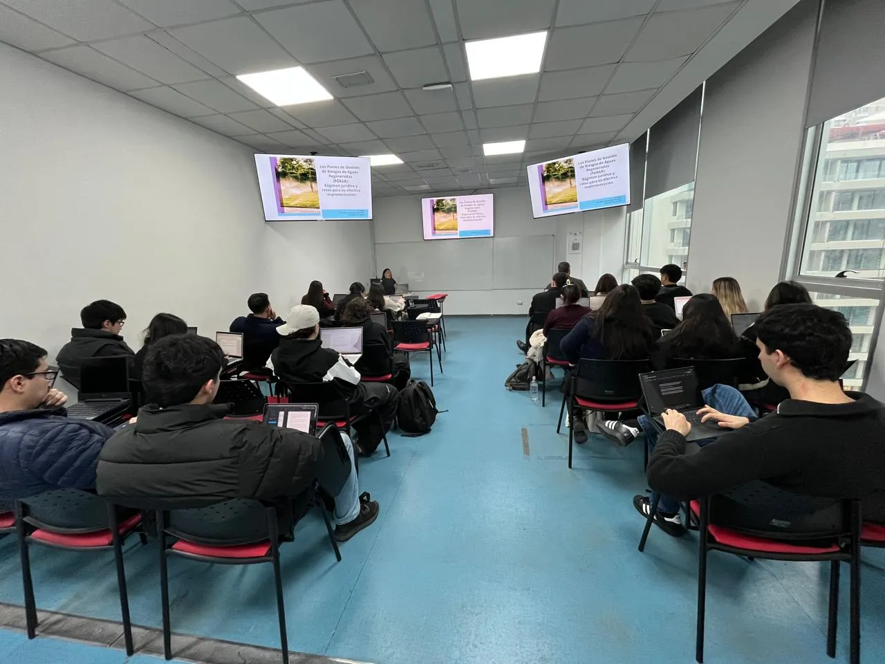 Students seated in a classroom with laptops open, facing a presenter at the front with slides on screens.