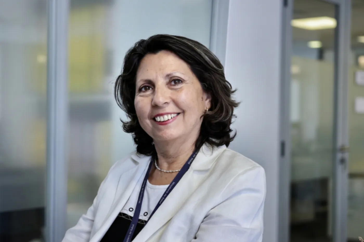 Smiling professional woman with dark hair in a white blazer and lanyard in an office setting