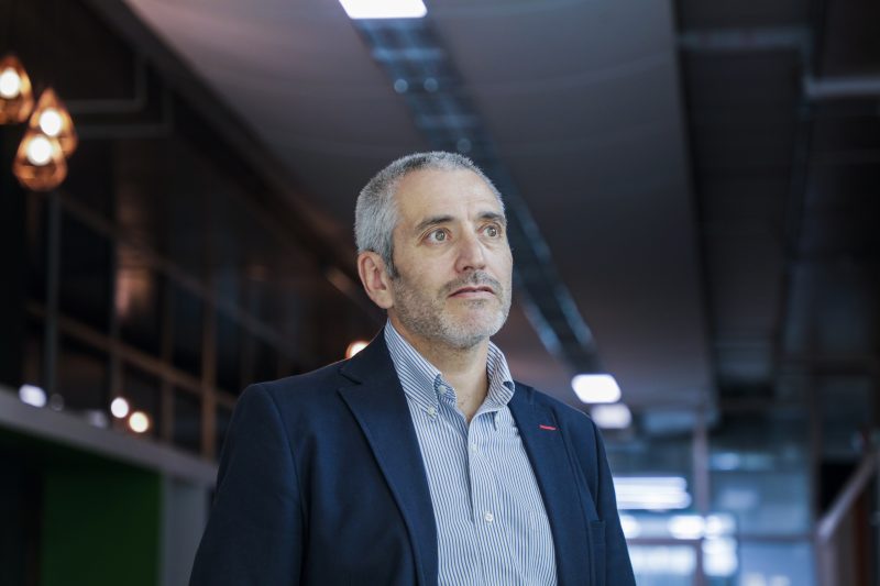 Man in a navy blazer and striped shirt stands in a modern office hallway, looking thoughtfully ahead.
