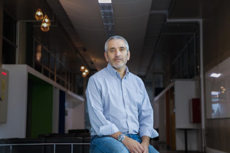 Middle-aged man with gray hair and beard sits on a bench in a modern office lobby, wearing a light blue striped shirt.