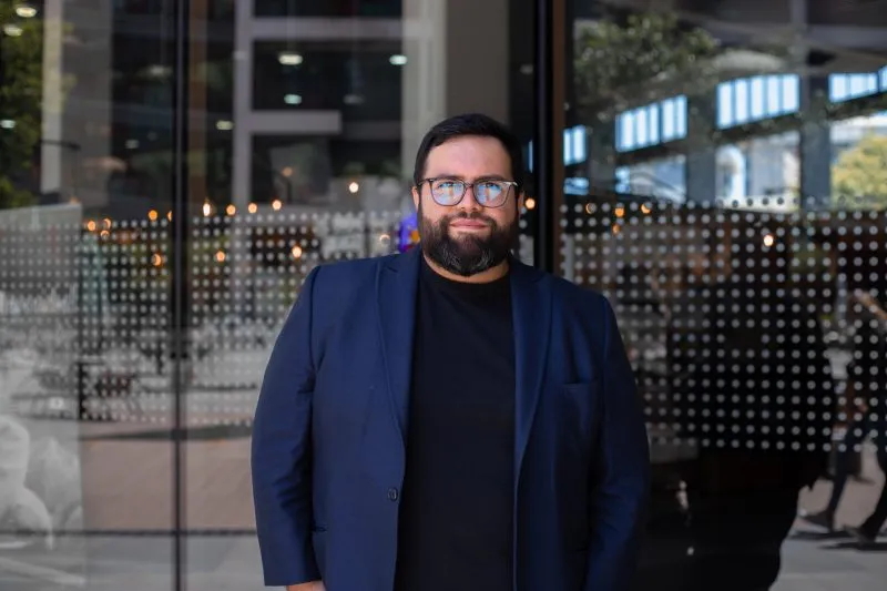 Man with dark hair and beard in a navy blazer and glasses stands outside a glass storefront.