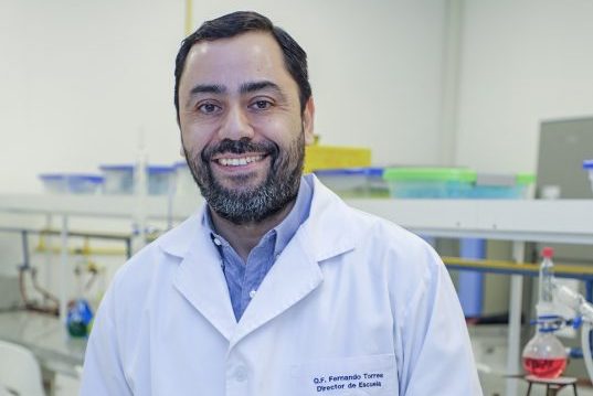 Smiling man in a white lab coat standing in a laboratory with benches and equipment in the background.