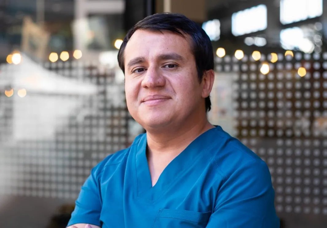 Healthcare professional in blue scrubs posing with arms crossed in front of a glass wall.
