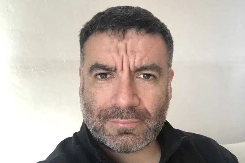 Close-up of a bearded man with dark hair frowning at the camera, wearing a black zippered top.