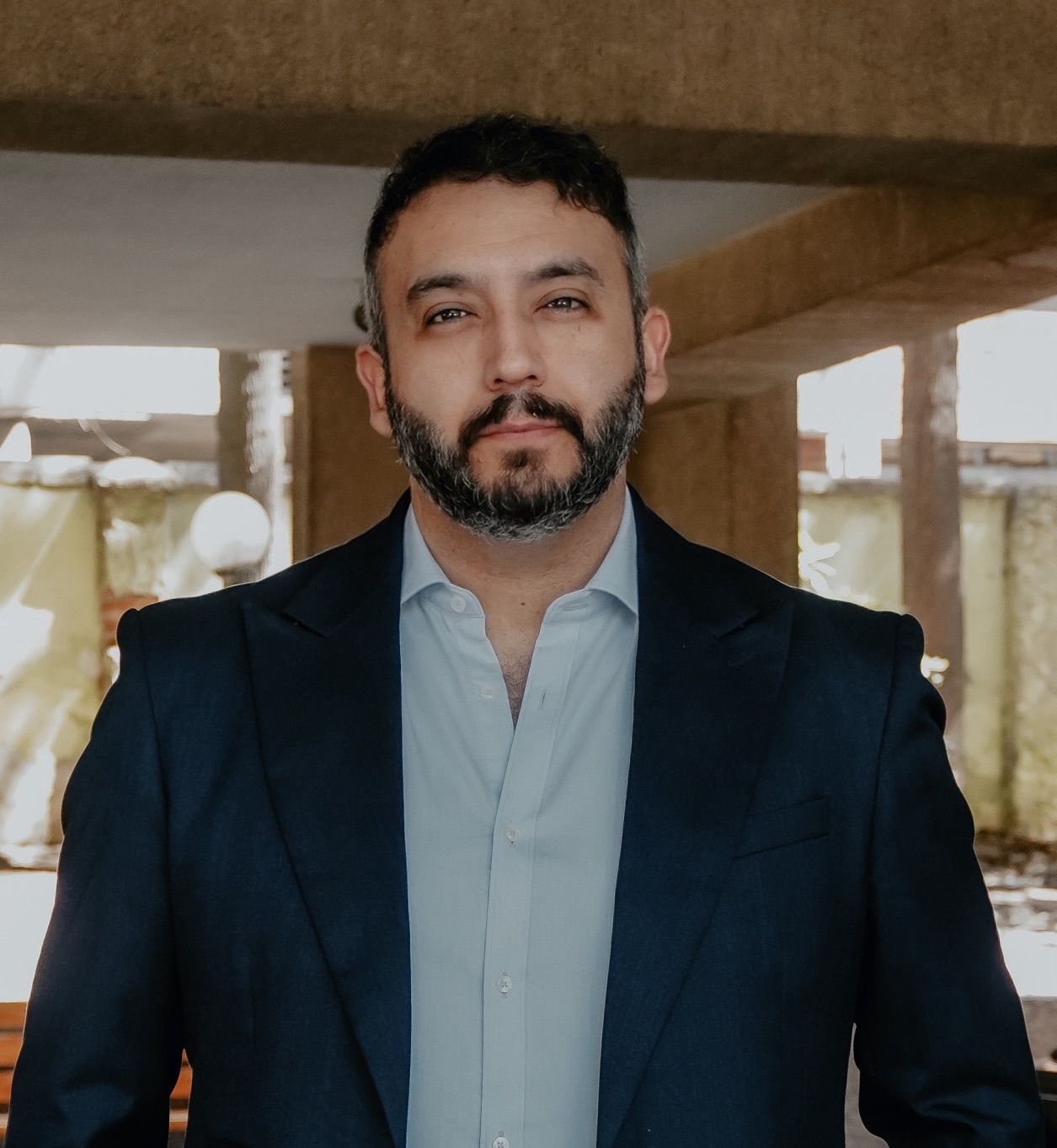 Head-and-shoulders portrait of a man with dark hair and beard, wearing a navy blazer and light blue shirt, standing in a sunlit concrete setting.