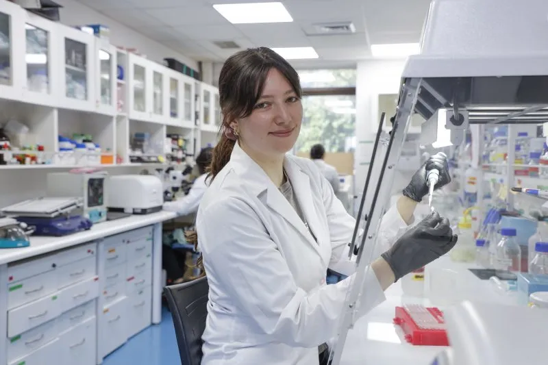 Female scientist in a white lab coat pipetting at a laboratory bench under a hood.