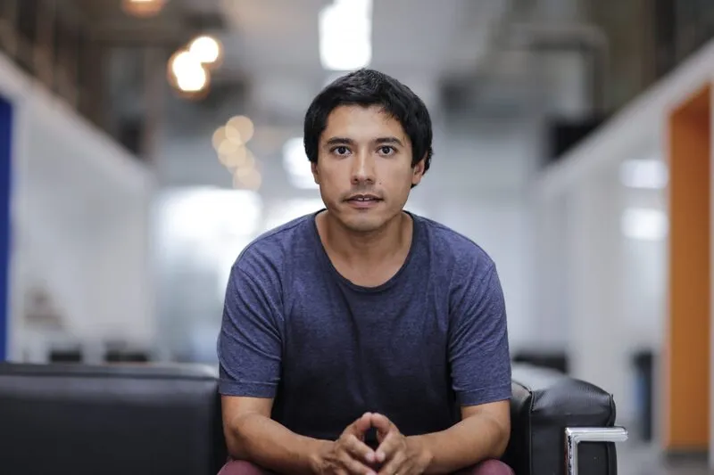 Young man in a blue t-shirt sits on a black couch in a bright, modern office setting and looks at the camera.