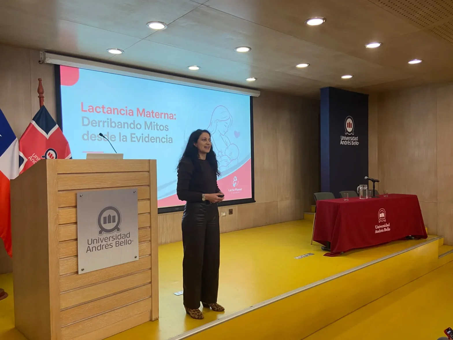 Speaker on a stage beside a lectern with 'Universidad Andrés Bello' sign, presentation slide behind reading 'Lactancia Materna: Derribando Mitos desde la Evidencia'
