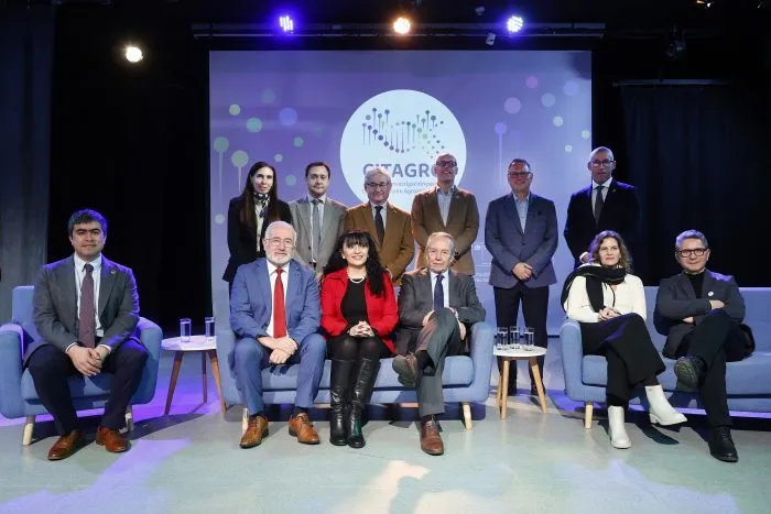 Group of professionals on a stage posing for a conference photo, some seated on blue chairs and others standing, with a projected logo behind them.