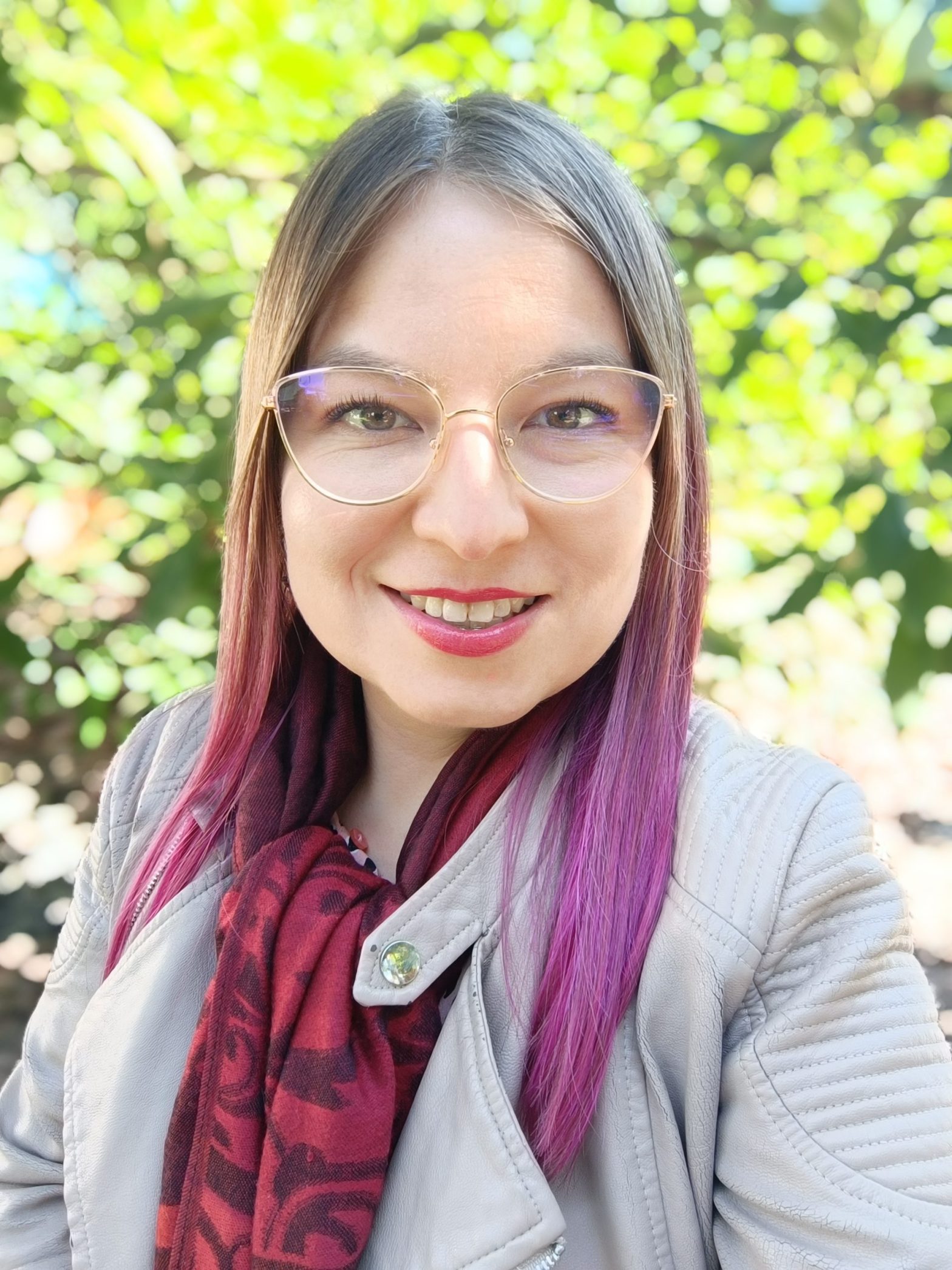 Close-up portrait of a smiling woman outdoors with round glasses, light jacket, and red scarf, green blurred foliage in the background.