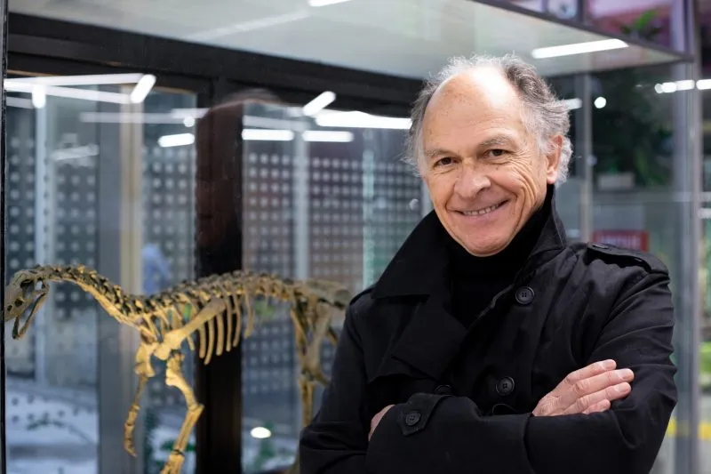 Smiling man in a black coat with arms crossed stands beside a golden dinosaur skeleton display in a museum.