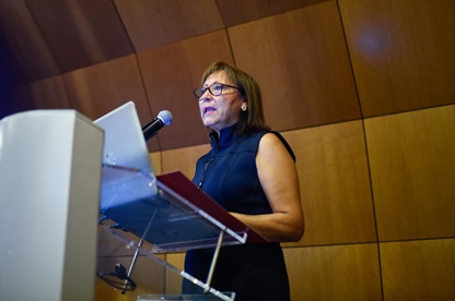 Woman wearing a dark sleeveless top speaks at a lectern with a microphone in a wood-paneled room.