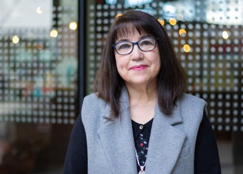 Portrait of a smiling woman with dark hair and glasses wearing a gray vest, standing outside a glass storefront.