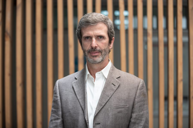 Man with salt-and-pepper hair and beard in a light brown blazer and white shirt, standing in front of vertical wooden slats.