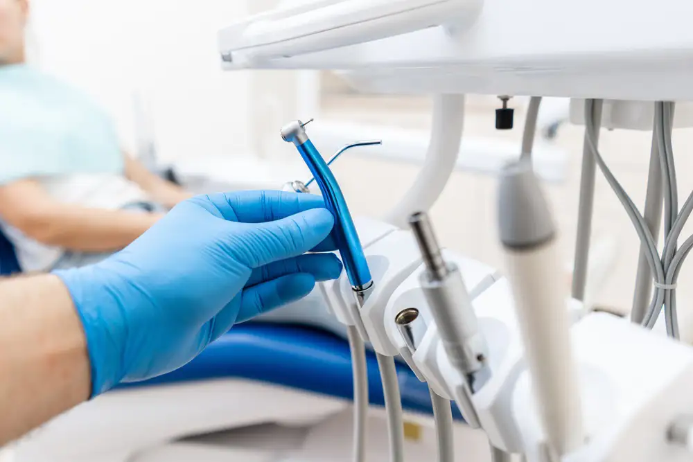 Dental clinician wearing blue gloves holds a blue dental instrument near a patient in a treatment chair.