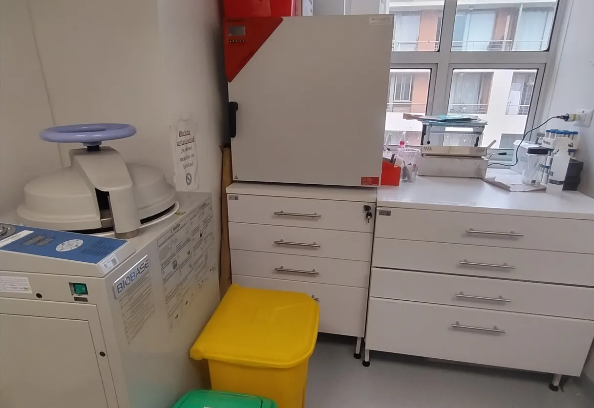 Laboratory workspace with a white benchtop, a BIOBASE centrifuge on the left, a tall white cabinet and drawers in the center, and yellow and green waste bins in the foreground near a windowed wall.